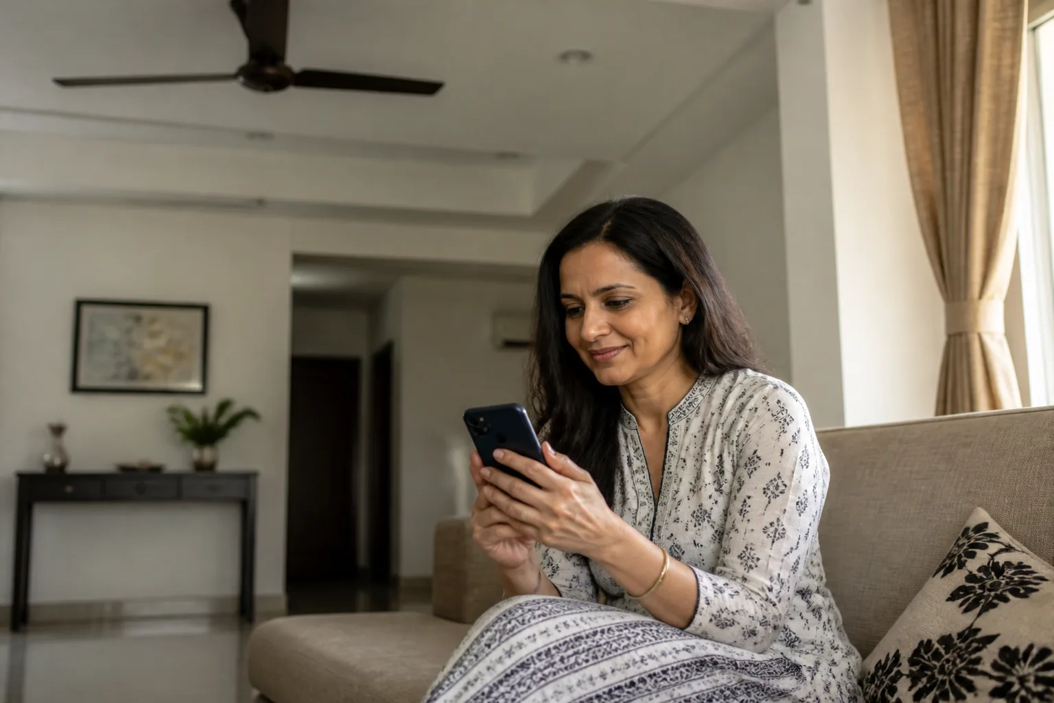 Indian woman in her 40s sitting on a sofa at home, looking at her phone in afternoon light